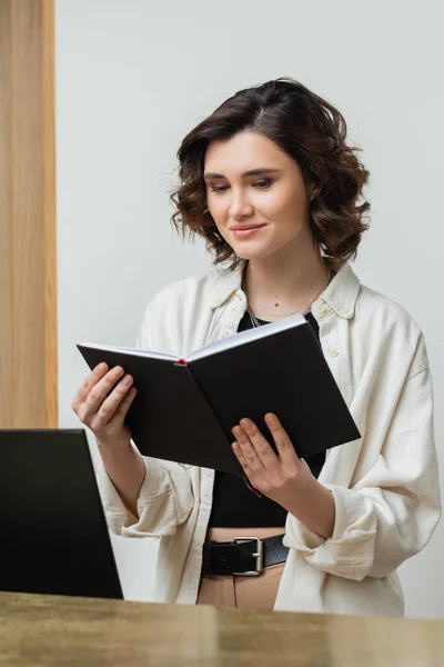 Cheerful concierge in trendy casual clothes and with wavy brunette hair looking at notebook near computer monitor while working at reception desk in contemporary hotel, digital nomad — Stock Photo