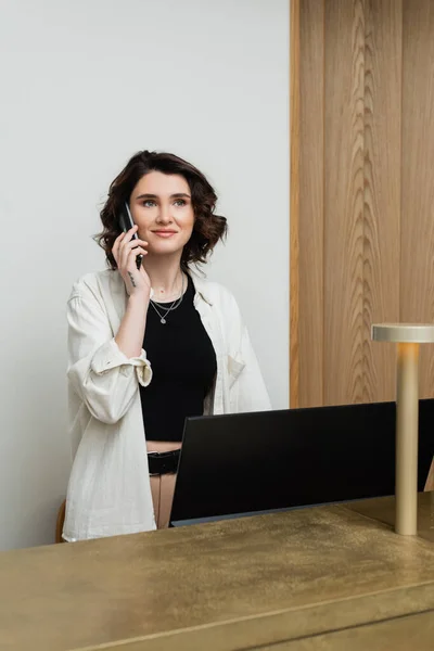 Happy concierge with wavy brunette hair and tattoo wearing stylish clothes and talking on telephone at reception desk near computer monitor and lamp in lobby of modern hotel, customer service — Stock Photo