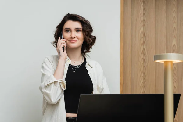 Pleased young woman in stylish casual clothes, with wavy brunette hair and tattoo talking on telephone while working as receptionist at front desk near computer monitor and lamp in hotel lobby — Stock Photo