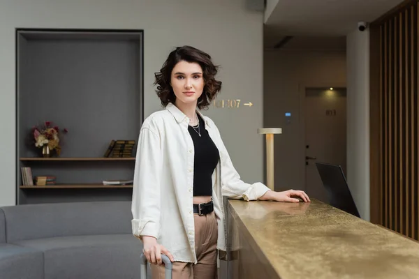 Confident woman in black crop top, white shirt and beige pants, with wavy brunette hair standing near lamp on reception desk and looking at camera in lobby of modern hotel, travel lifestyle — Stock Photo