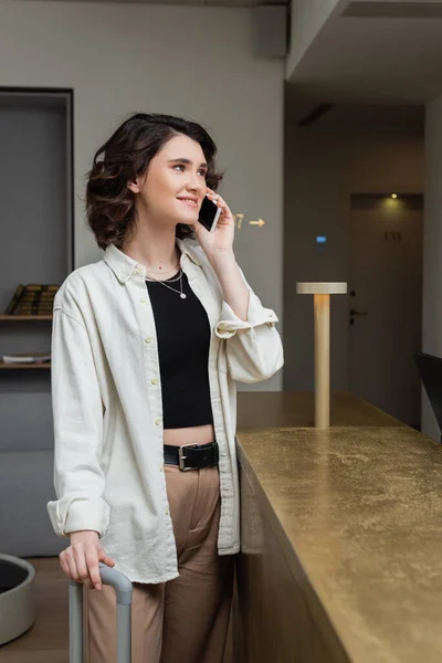 Carefree woman with wavy brunette hair, in white stylish shirt, black crop top and beige pants smiling and talking on mobile phone near front desk in lobby of modern hotel — Stock Photo