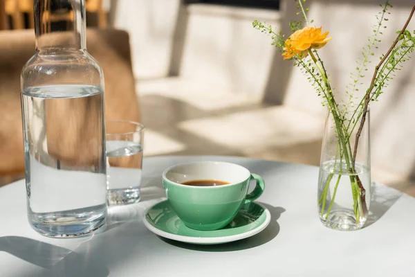 Vase with yellow rose and green plants, glass and decanter with fresh pure water, saucer, cup with black coffee on white round table in morning sunlight, outdoor terrace of hotel cafe — Stock Photo