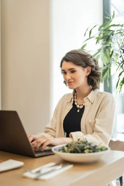 Positive woman with wavy brunette hair, in stylish casual clothes typing on laptop near fresh vegetable salad while sitting at table in lobby cafe of contemporary hotel, work and travel — Stock Photo