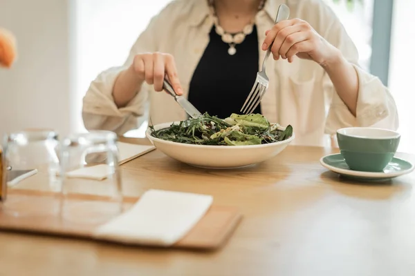 Partial view of young woman with fork and knife eating fresh vegetable salad near cup of aromatic coffee, saucer, glasses and napkins on blurred foreground on table of lobby cafe in modern hotel — Stock Photo
