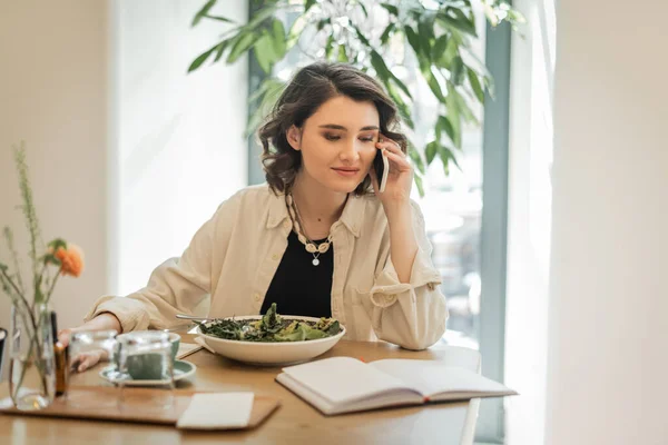 Positive woman in stylish casual clothes looking at notebook during conversation on mobile phone near fresh vegetable salad, glasses and cup of aromatic coffee in hotel lobby, work-life integration — Stock Photo