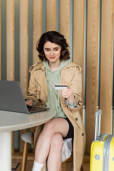 Atraente jovem mulher em elegante casaco bege trincheira e com cabelo morena ondulado sentado à mesa com laptop e segurando cartão de crédito durante compras na internet no lobby do hotel contemporâneo — Fotografia de Stock