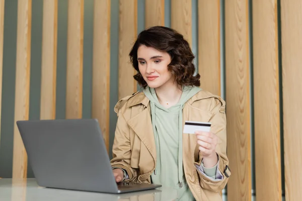 Pleased woman with wavy brunette hair, in beige trench coat holding credit card and looking at laptop while sitting at table in lobby of contemporary hotel during online shopping, leisure and travel — Stock Photo