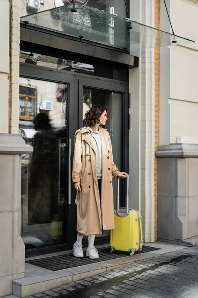 Junge Frau in beigem Trenchcoat, grauem Kapuzenpulli und weißen Turnschuhen mit gelber Reisetasche am Eingang eines modernen Hotels in einer europäischen Stadt, Reiselebensstil — Stockfoto
