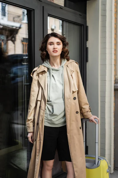 Confident woman with wavy brunette hair, in grey hoodie and beige trench coat standing with yellow suitcase and looking at camera near glass doors of modern hotel on city street, travel lifestyle — Stock Photo