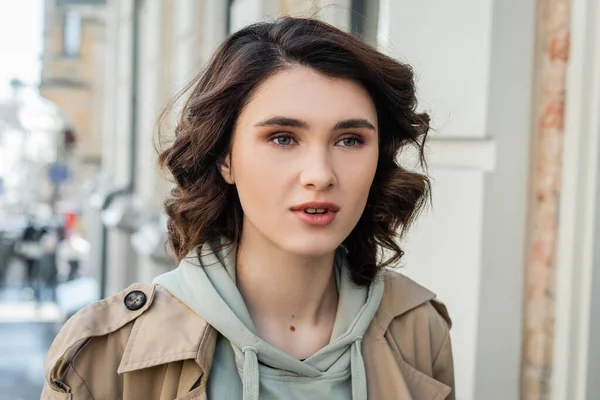 Portrait of charming and fashionable woman with short brunette hair, in beige trench coat and grey hoodie looking away in European city on blurred background, street photography — Stock Photo