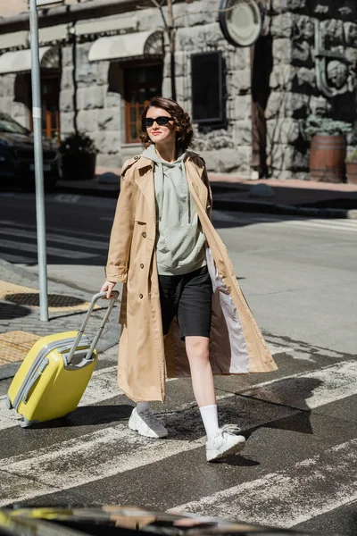 Full length of self-assured and independent woman in beige trench coat, grey hoodie and black shorts holding yellow suitcase while crossing road in European city, urban lifestyle — Stock Photo