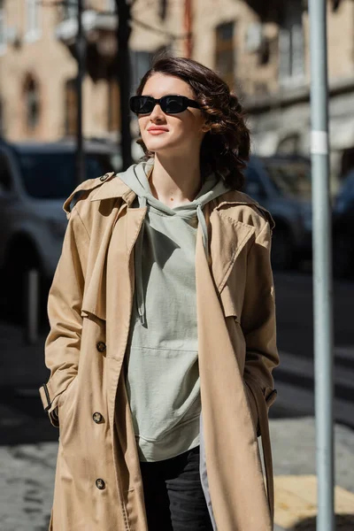Молодая и позитивная женщина с волнистыми волосами брюнетки, в темных солнцезащитных очках, серой толстовке и бежевом плаще, смотрящая в сторону на размытом фоне в европейском городе, уличная фотография — стоковое фото
