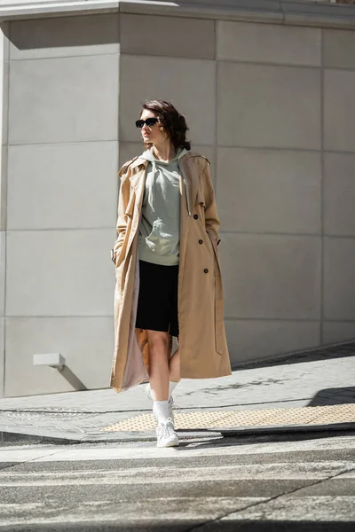 Full length of trendy woman with wavy brunette hair crossing road in beige trench coat, grey hoodie, black shorts, white sneakers and dark sunglasses near modern building in city — Stock Photo