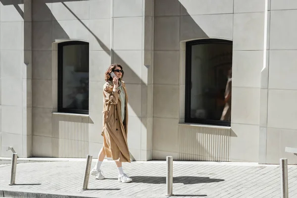 Eine junge, modische Frau in dunkler Sonnenbrille, beigem Trenchcoat und weißen Turnschuhen, die mit dem Smartphone spricht, während sie durch ein modernes Gebäude auf der Straße in der europäischen Stadt läuft — Stockfoto