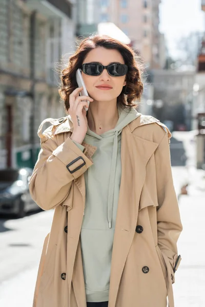 Young tattooed woman with wavy brunette hair, in dark sunglasses, beige trench coat and grey hoodie standing and talking on mobile phone on urban street on blurred background — Stock Photo