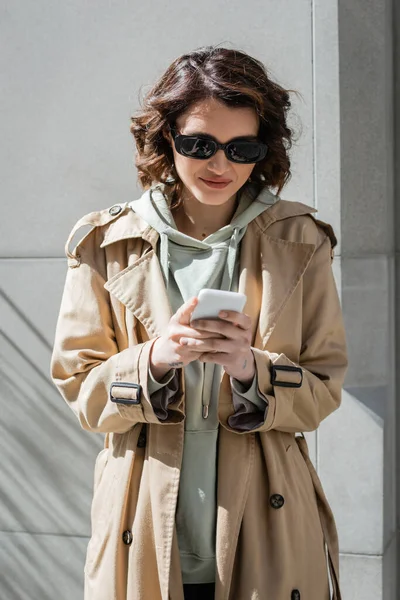 Young and carefree woman with wavy brunette hair and tattoo, in dark sunglasses, beige trench coat and hoodie browsing internet on mobile phone while standing near grey wall in sunlight on street — Stock Photo