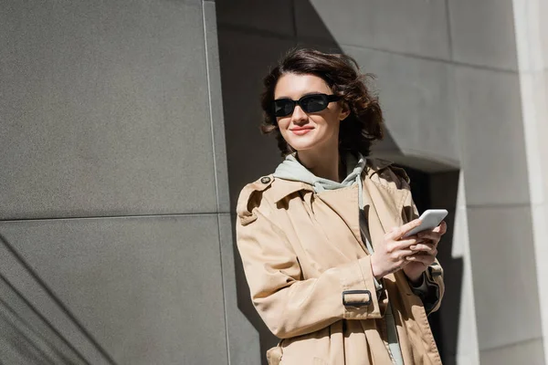 Pleased and trendy woman in dark sunglasses, hoodie and beige trench coat messaging on smartphone and looking away while standing near grey building in city, urban lifestyle, street photography — Stock Photo