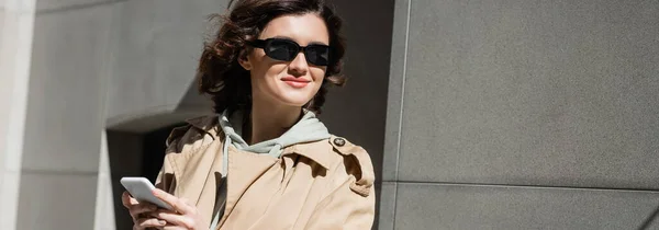 Mujer encantada y elegante en gafas de sol oscuras, gabardina beige y sudadera con capucha en el teléfono móvil mientras está de pie cerca del edificio gris en la calle urbana de la ciudad europea, pancarta - foto de stock