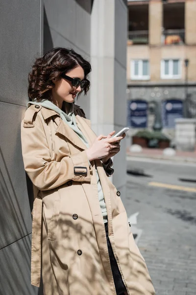 Cheerful and trendy woman with wavy brunette hair and tattoo, in dark sunglasses and beige trench coat standing near grey building and browsing internet on mobile phone on street in european city — Stock Photo