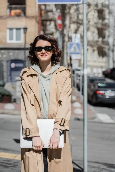 Carefree woman with wavy brunette hair and tattoo, wearing beige trench coat, grey hoodie and dark sunglasses while standing with laptop on urban street of european city on blurred background — Stock Photo