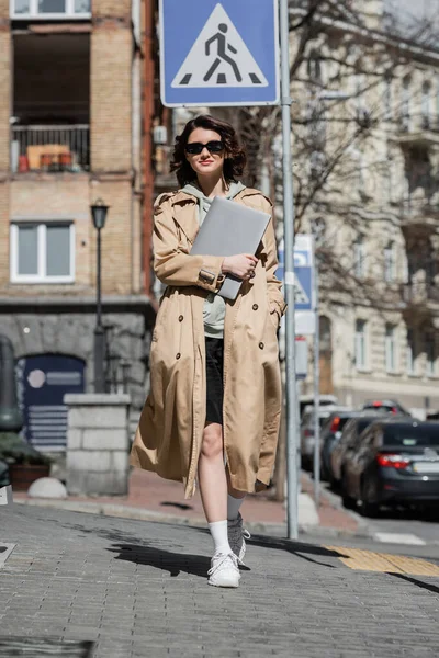 Young and stylish woman with wavy brunette hair, in dark sunglasses, beige trench coat and white sneakers walking with laptop on sunny street of European city, work and travel, freelance lifestyle — Stock Photo