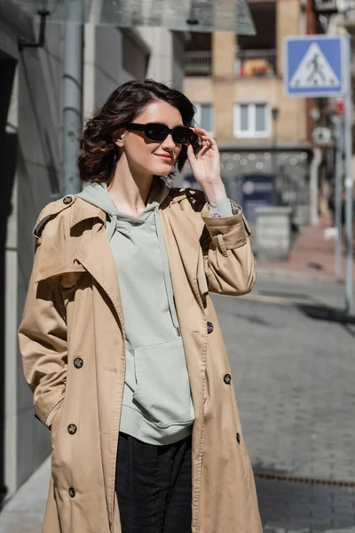 Young appealing woman with wavy brunette hair adjusting dark stylish sunglasses while standing in grey hoodie and beige trench coat in city on blurred background, street photography — Stock Photo