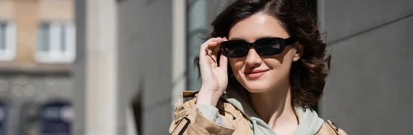 Portrait of young and joyful woman with wavy brunette hair, wearing beige trench coat and grey hoodie, adjusting dark stylish sunglasses and looking away on urban street, banner — Stock Photo