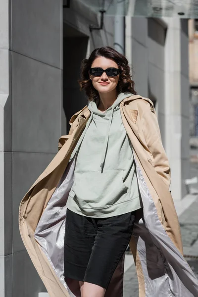 Stylish and carefree woman in dark sunglasses, grey hoodie and beige trench coat posing on street in modern European city and looking at camera, travel lifestyle, urban fashion — Stock Photo