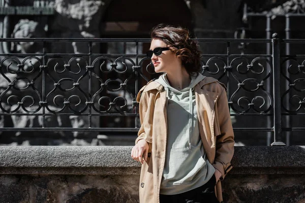 Travel lifestyle, young and fashionable woman with wavy brunette hair, in dark sunglasses, beige trench coat and grey hoodie looking away while standing near forged fence on city street, travel lifestyle — Stock Photo