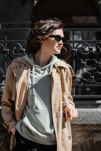 Young and confident woman in grey hoodie, beige trench coat and dark stylish sunglasses standing with hand in pocket and looking away near forged fence on city street — Stock Photo