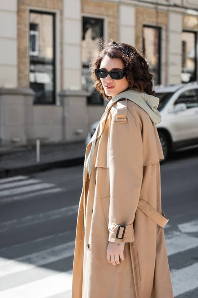Привлекательная женщина с волнистыми брюнетками гуляет в темных стильных солнцезащитных очках, серой толстовке и бежевом плаще, глядя на камеру на размытой улице в европейском городе, городская мода — стоковое фото
