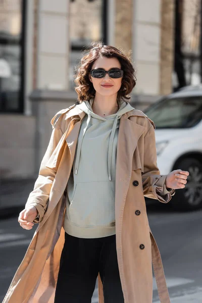 Стильная и улыбающаяся женщина в темных стильных солнцезащитных очках, серой толстовке и бежевом плаще, смотрящая на камеру во время прогулки по улице европейского города на размытом фоне — стоковое фото