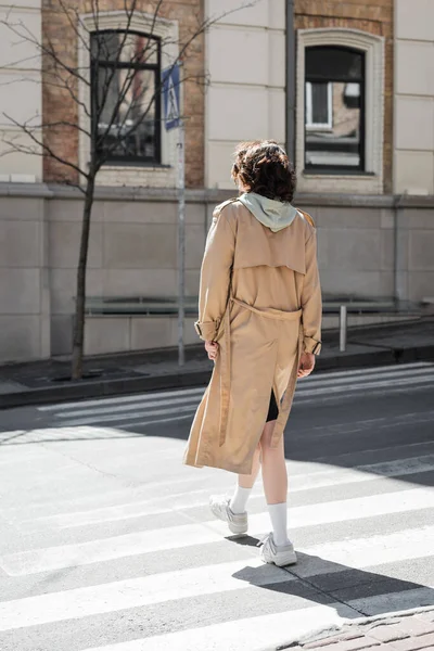 Full length of young woman with wavy brunette hair, in beige trench coat, grey hoodie and white sneakers crossing road on sunny street of European city, back view, urban fashion, travel lifestyle — Stock Photo
