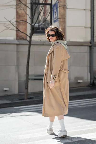 Full length of young and stylish woman with wavy brunette hair crossing road in dark sunglasses, grey hoodie, beige trench coat and white sneakers and looking at camera in European city — Stock Photo