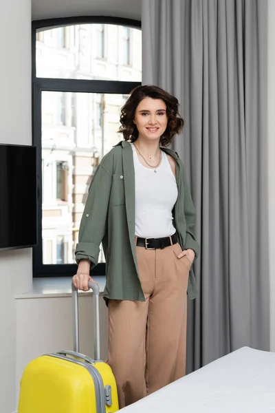 Smiling woman with wavy brunette hair and in stylish clothes looking at camera while standing with hand in pocket near window, grey curtains, lcd tv and bed in cozy hotel suite — Stock Photo