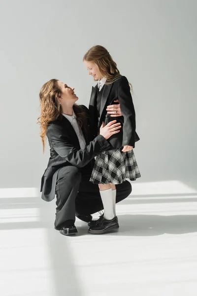 Щаслива мати і дочка, бізнес-леді в костюмі обіймають школярку в уніформі з плетеною спідницею, блейзери, готуються до нового навчального року, заохочуючи, дивлячись один на одного — стокове фото