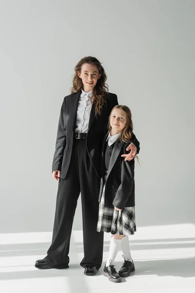 Modern mother and daughter, businesswoman in suit hugging schoolgirl in uniform with plaid skirt, on grey background, blazers, getting ready for new school year, looking at camera, formal attire — Stock Photo