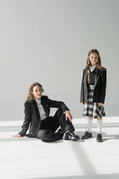 Working mother and child, businesswoman in suit sitting near schoolgirl in uniform with plaid skirt on grey background, blazers, new school year, looking at camera, formal attire — Stock Photo