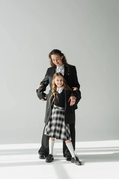 Crianza moderna, mujer de negocios de moda en traje abrazando a la hija en uniforme escolar con falda a cuadros y de pie juntos sobre fondo gris, madre feliz y el niño - foto de stock