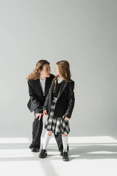 Schoolgirl looking at working mother, cheerful girl in school uniform standing with businesswoman in suit on grey background, formal attire, fashionable family, bonding, modern parenting — Stock Photo