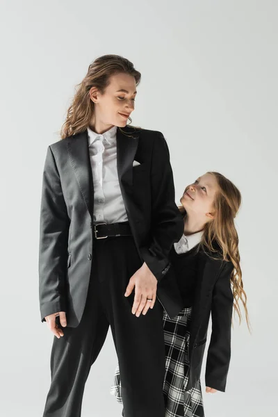 Estudante olhando para sua mãe, menina alegre em uniforme escolar de pé com mulher de negócios em terno em fundo cinza, traje formal, família na moda, colagem, parentalidade moderna — Fotografia de Stock