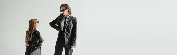 Modern mother and daughter in sunglasses, businesswoman in suit and schoolgirl in uniform looking at each other while standing on grey background in studio, fashionable family, banner — Stock Photo