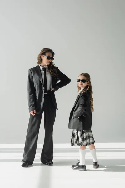 Весела жінка зі своєю дочкою, бізнес-леді в костюмі позує з рукою на стегно і школярка в сонцезахисних окулярах і форма, що стоїть разом на сірому фоні в студії, офіційний одяг — стокове фото