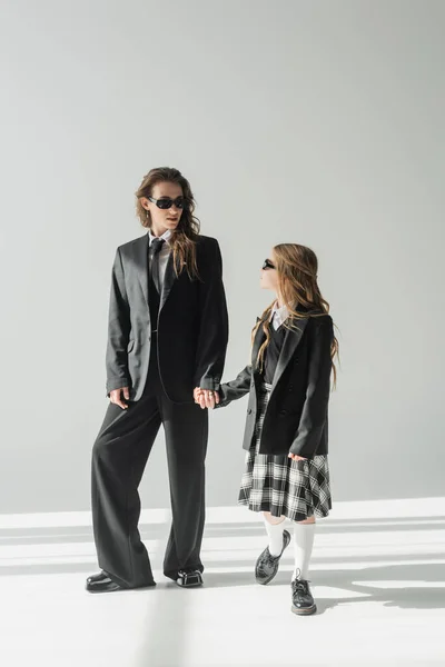 Stylish mother and child holding hands, businesswoman in formal attire and sunglasses looking at schoolgirl in uniform on grey background in studio, fashionable family — Stock Photo