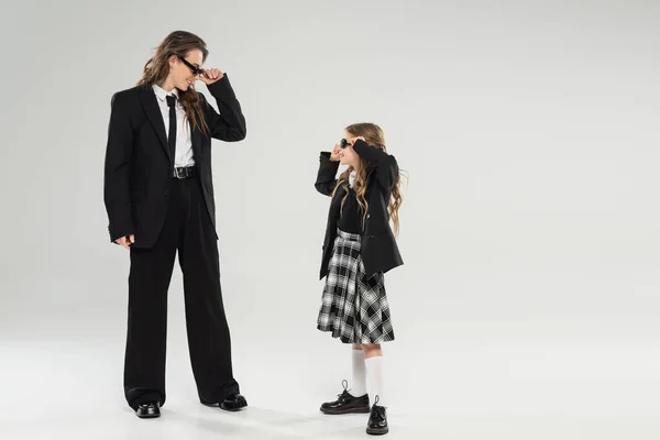 Стильна мати і дитина в сонцезахисних окулярах, щаслива бізнес-леді в офіційному вбранні, дивлячись на школярку в уніформі на сірому фоні в студії, модна сім'я, сучасне батьківство — стокове фото