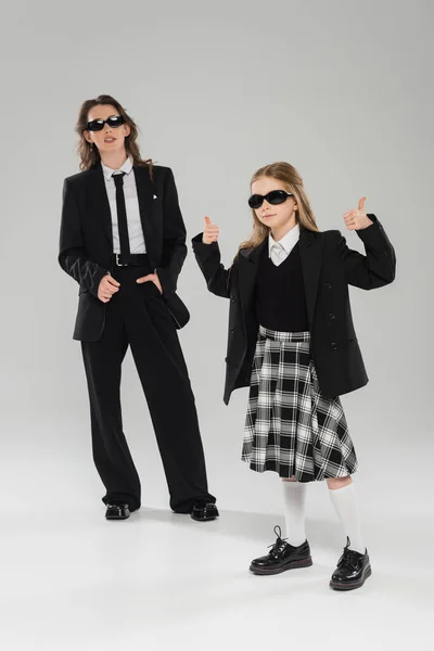 Stylish schoolgirl in sunglasses and uniform showing thumbs up and standing near modern mother on grey blurred background in studio, formal attire, back to school — Stock Photo