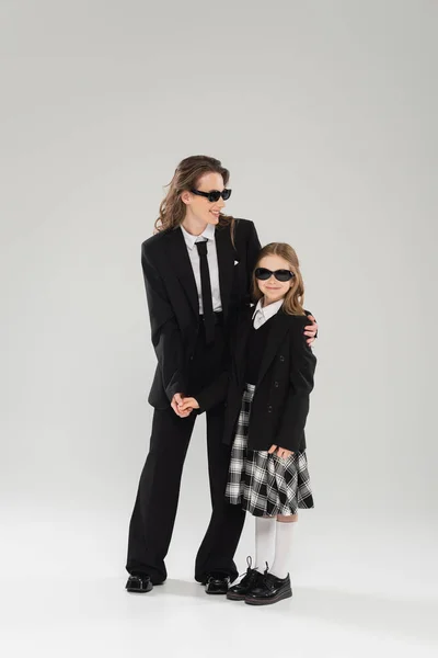 Crianza moderna, mujer de negocios feliz en traje y gafas de sol abrazando a la hija en uniforme escolar con falda a cuadros y de pie juntos sobre fondo gris, madre e hijo - foto de stock