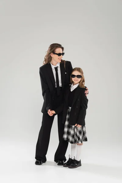 Parent moderne, femme d'affaires heureuse en costume et lunettes de soleil étreignant fille en uniforme scolaire avec jupe à carreaux et debout ensemble sur fond gris, mère et enfant — Photo de stock