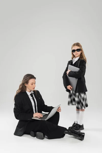 Digital nomadism, fashionable woman in suit using laptop near daughter in school uniform and sunglasses on grey background, remote work, working mother, business attire — Stock Photo