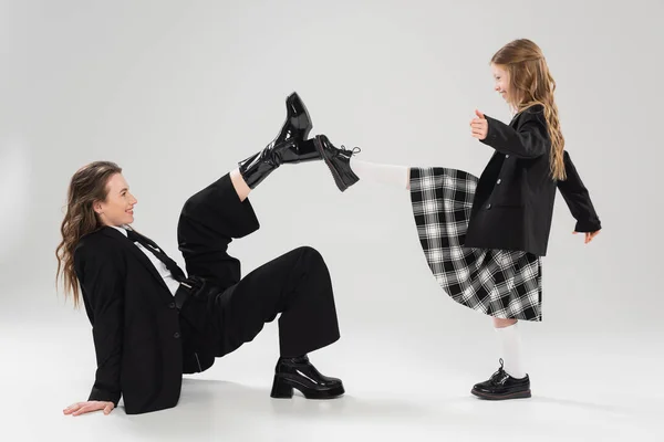Modern parenting, having fun, side view of happy mother in suit and child in school uniform touching boots of each other on grey background in studio, blazer, businesswoman, back to school — Stock Photo